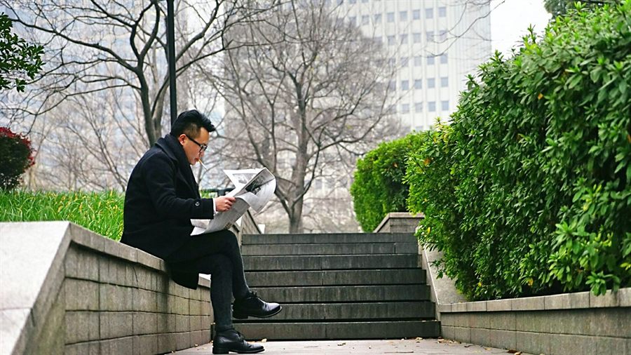 Man in a suit reading a newspaper on a low wall in a city park